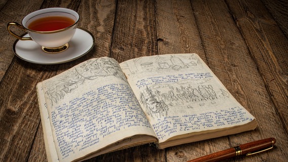 Neben einem aufgeschlagenen alten, handschriftlich beschriebenem Buch steht eine Tasse Tee. © imago images / Zoonar 