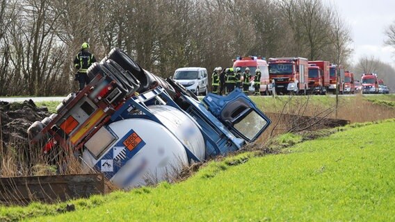 Ein Tanklaster liegt umgekippt in einem Graben, Feuerwehrleute und Fahrzeuge stehen auf der angrenzenden Bundesstraße. © Westküstennews Foto: Florian Sprenger