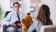 Eine Frau mit einem Teddybär im Ar sitzt auf einer Couch und spricht und ihr gegenüber sitzt ein man und macht sich Notizen. © imago images / Zoonar II 