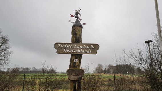 An einem Pfahl in einer ländlichen Gegend hängt ein Schild mit der Aufschrift "Tiefste Landstelle Deutschlands". © NDR Foto: Lisa Pandelaki