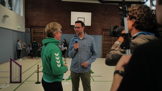 NDR-Moderator Christopher Scheffelmeier interviewt jemanden in einer Turnhalle © NDR Foto: Lisa Pandelaki