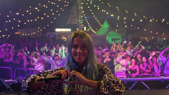 DJ 2Elements formt ein Herz mit ihren Händen und lächelt in die Kamera. Im Hintergrund ist das Publikum zu sehen. © NDR Foto: Christian Wolf