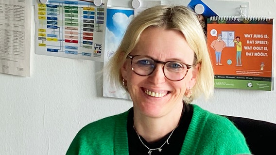 Eine Frau mit schulterlangen blonden Haaren und einer grünen Cardigan sitzt in einem Büro und lächelt in die Kamera. © NDR Foto: Jan Graf