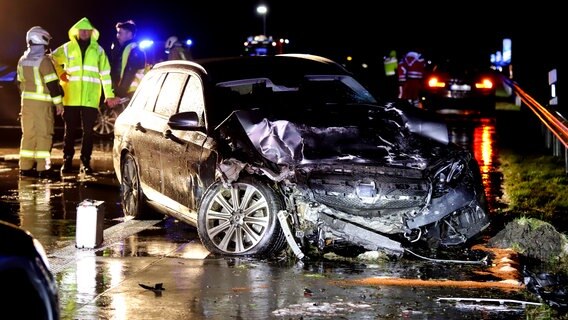 Ein Auto steht mit zerstörter Front entgegen der Fahrtrichtung an einer Leitplanke der A23 bei Tensbüttel-Röst, im Hintergrund stehen Rettungskräfte der Feuerwehr. © Westküstennews Foto: Florian Sprenger