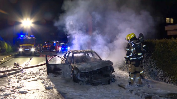 Ein ausgebranntes Fahrzeugt steht umgeben von Löschschaum und Feuerwehr auf der Straße. © TV News Kontor Foto: Screenshot