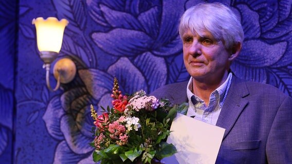 Wolfgang Krischke steht mit einem Blumenstrauß und einer Urkunde auf der Bühne im Ohnsorg Theater in Hamburg beim "Vertell doch mal". © NDR Foto: Oke Jens