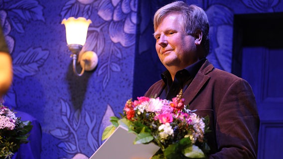 Heiko Thomsen steht mit einem Blumenstrauß und einer Urkunde auf der Bühne im Ohnsorg Theater in Hamburg beim "Vertell doch mal". © NDR Foto: Oke Jens