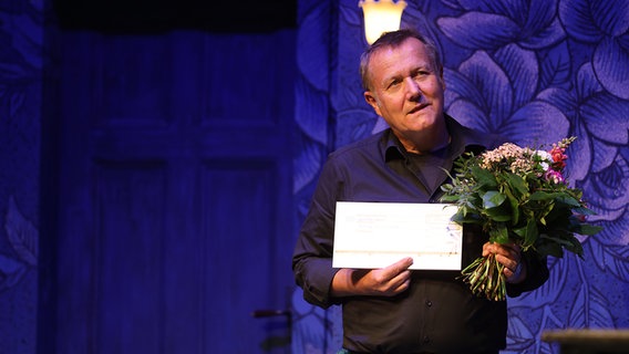 Holger Wittschen steht mit einem Blumenstrauß und einer Urkunde auf der Bühne im Ohnsorg Theater in Hamburg beim "Vertell doch mal". © NDR Foto: Oke Jens