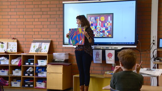 Eine Lehrerin steht vor einer Schulklasse und hält ein Kunstwerk in Händen. © NDR 