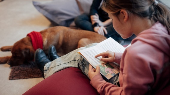 Ein Mädchen liest in einem Buch und zu ihren Füßen liegt ein Hund. © NDR Foto: Lisa Pandelaki