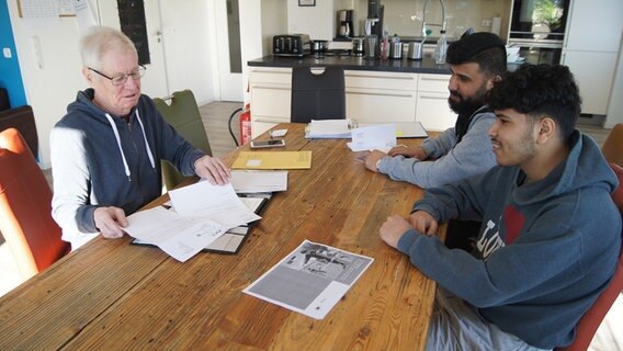 Links Edgar Manthei, sitzt gegenüber von Muhammed und seinem großen Bruder an einem Holztisch auf dem Papiere ausgebreitet liegen. © NDR Foto: Astrid Wulf