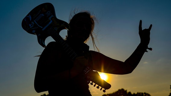 Eine Frau mit E-Gitarre auf der Schulter zeigt auf dem Wacken-Festivalgelände den Metalgruß. © dpa-Bildfunk Foto: Axel Heimken