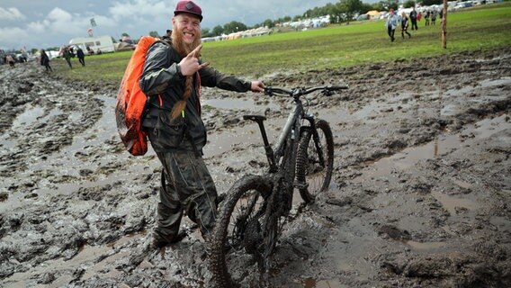 Metal-Fan Lukas Litwin aus Bochum ist am 1. August 2023 mit seinem E-Mountainbike auf dem schlammigen Festivalgelände von Wacken unterwegs. © picture alliance/dpa Foto: Christian Charisius