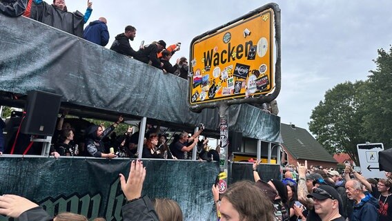 Ein Bus fährt an dem Ortsschild von Wacken vorbei. Mehrere Fans des Wacken Open Air säumen die Straße. © NDR Foto: Jörn Zahlmann