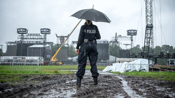 Eine Person steht mit Regenschirm und Gummistiefeln im Schlamm auf dem Wacken Open Air. Auf der Rückseite ihres Overalls steht: Polizei. © NDR Foto: Katrin Juister-Ahlfeld