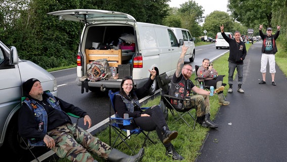Metalfans sitzen am Rande eines Staus auf Campingstühlen und zeigen den Metalgruß. © Christian Charisius/dpa 