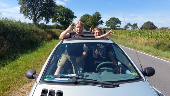 Zwei Männer lehnen sich für ein Foto aus dem offenen Cabriodach, während sie bei der Anreise zum Wacken-Festival im Stau stehen © NDR Foto: Laura Albus