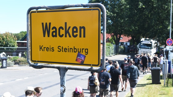 Wacken Besucher laufen an dem Dorfschild "Wacken Kreis Steinburg" durch die Straßen © NDR Foto: Jörn Zahlmann