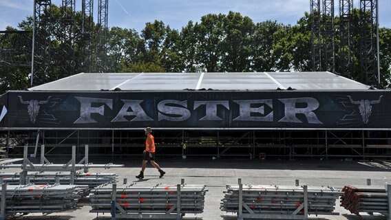 Ein Bauarbeiter in einer gelben Sicherheitsweste läuft an einer Bühne auf dem Festival-Gelände mit dem schwarz-weißen Banner "FASTER" vorbei. © NDR Foto: Eva Schulze-Gabrechten