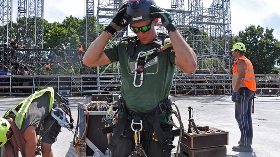 Bauarbeiter rüsten sich mit Schutzausrüstung für den Aufbau des Schädels auf dem Festival-Gelände in Wacken aus. © NDR Foto: Eva Schulze-Gabrechten