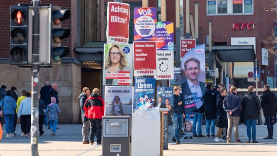 Wahlplakate verschiedener Parteien hängen an Straßenlaternen teilweise übereinander. Rundherum stehen Passanten.  Foto: penofoto