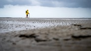 Ein Mann in einer gelben Jacke spaziert unter bewölktem Himmel durch das Watt (Themenbild). © picture alliance/dpa | Sina Schuldt Foto: Sina Schuldt