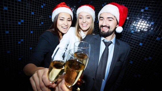 Drei Personen mit Weihnachtsmützen auf stoßen mit Sekt an. © imago images 