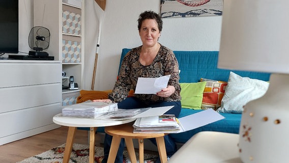 Eine Frau sitzt in ihrem Wohnzimmer mit einem Blatt Papier in der Hand und blickt in die Kamera. © NDR Foto: Hannah Böhme