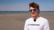 Ein Mann mit Sonnenbrille steht am Strand und lächelt in die Kamera. © NDR 