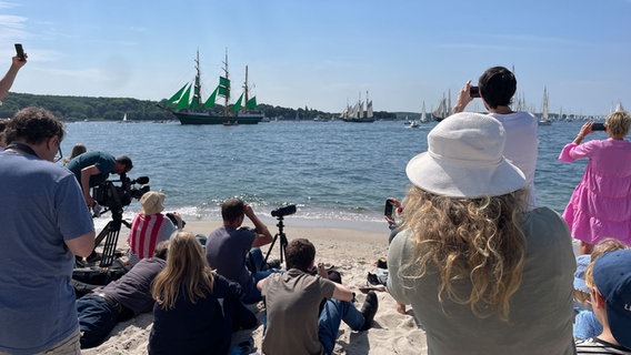 Zahlreiche Menschen tummeln sich an einem Strandabschnitt, um die Windjammerparade 2023 in Kiel zu beobachten. © NDR Foto: Paul Wessel