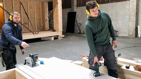 Zwei Männer bauen ein Bodenelement in einer Werkshalle zusammen. © NDR 