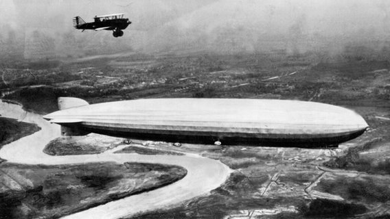 Ein Zeppelin fliegt 1928 über eine Landschaft begleitet von einem Doppeldeckerflugzeug. © dpa picture-alliance 