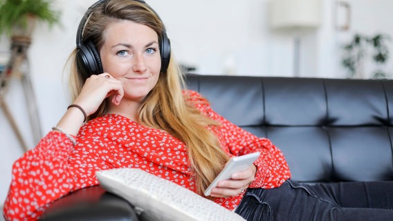 Eine junge Frau mit Kopfhörern sitzt auf dem Sofa und hält ein Handy. © photocase Foto: 2Design