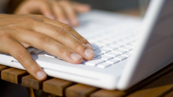 Zwei Hände betätigen eine Tastatur auf einem weißen Laptop, der im sonnigen Licht auf einem Holztisch liegt. © photocase Foto: soulcore