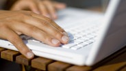 Zwei Hände betätigen eine Tastatur auf einem weißen Laptop, der im sonnigen Licht auf einem Holztisch liegt. © photocase Foto: soulcore