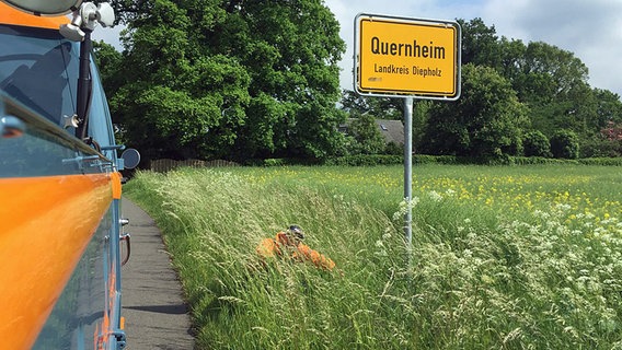 Schüssel-Schorse kniet neben dem Ortsschild von Quernheim im Gras. © NDR Foto: Andi Gervelmeyer