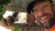 Schüssel-Schorse sitzt in einem Wagen, während eine Giraffe ihre Kopf durch die Seitenscheibe steckt. © NDR 