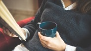 Eine junge Frau im blauen Pullover sitzt gemütlich im Sessel und hält einen Kaffeebecher und ein Buch in ihren Händen. © photocase.com Foto: Christine ten Winkel