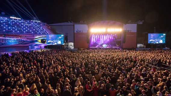 Publikum und Bühne beim NDR 2 Papenburg Festival, links im Hintergrund liegt ein farbig angestrahltes Kreuzfahrtschiff © NDR 2 Foto: Axel Herzig