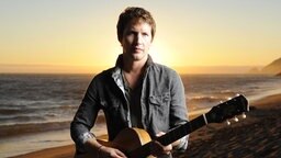 Der britische Sänger James Blunt mit Gitarre am Strand bei Sonnenuntergang © Warner Music Group 