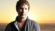 Der britische Sänger James Blunt mit Gitarre am Strand bei Sonnenuntergang © Warner Music Group 