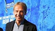 Jürgen Klinsmann lächelt bei seiner Vorstellung als Hertha-Trainer am 28. November 2019 in die Kamera, im Hintergrund das Vereinslogo. © imago images/Bernd König 