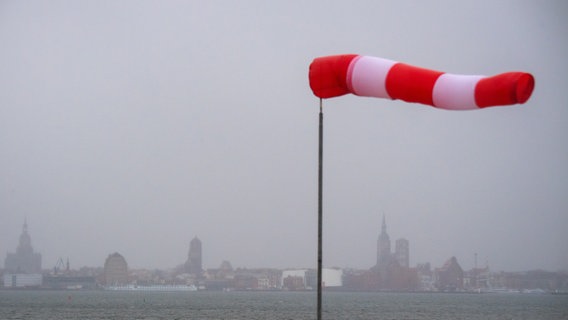 Ein Windsack steht fast horizontal in der Luft und zeigt den recht starken Wind an. Im Hintergrund ist die Silhouette der Hansestadt Stralsund zu sehen. In den kommenden Tagen wird stürmisches und regnerisches Wetter in Deutschland erwartet.  Foto: Stefan Sauer