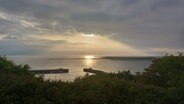 Dichte, dunkle Wolken überdecken die aufgehende Sonne am Himmel über Helgoland. In der Nordsee spiegelt sich ihr Licht. Im Vordergrund: Gebüsch.  