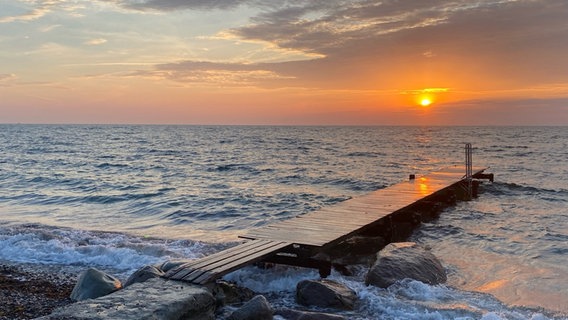 Die Sonne geht über der Ostsee auf, färbt das Wasser in orange-violettes Licht. Im Vordergrund ragt ein Steg über die ruhige See. © Privat Foto: NDR 2 Hörerinnen Brigitte und Inge