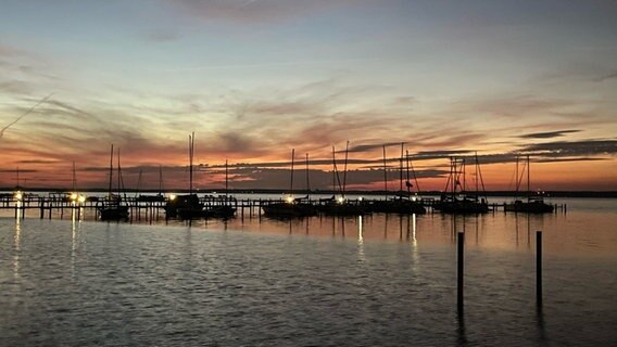 Die Morgensonne färbt den Himmel über dem Steinhuder Meer in orangefarbenes Licht. Im Vordergrund schwimmen Segelschiffe an einem Steg. © Privat Foto: NDR 2 Hörerin Monika