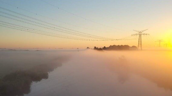 Ein Fluss fließt unter einer Stromtrasse hindurch. Am Horizont geht die Morgensonne auf und bahnt sich ihren Weg durch dichte Nebelschwaden.
färbt den Himmel über dem Steinhuder Meer in orangefarbenes Licht. Im Vordergrund schwimmen Segelschiffe an einem Steg. © Privat Foto: NDR 2 Hörer Tido
