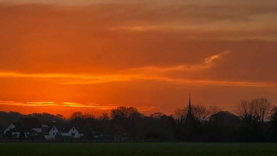 Die Sonne geht am Horizont auf, vor dem rot gefärbten Himmel sind Umrisse eines kleinen Ortes zu erkennen (Dedensen). © Privat Foto: NDR 2 Hörer Elke Schroth