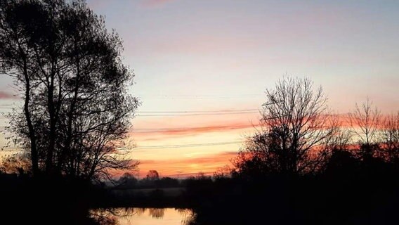Die Sonne geht am Horizont auf. Der orange-rot gefärbte Himmel spiegelt sich in einem Flusslauf. © Privat Foto: NDR 2 Hörerin Angelika