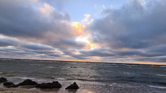 Die Sonne schaut durch die Wolkenlöcher am Himmel über der Ostsee. Ihre Strahlen färben den Himmel orange-blau ein. © Privat Foto: NDR 2 Hörer Didi
